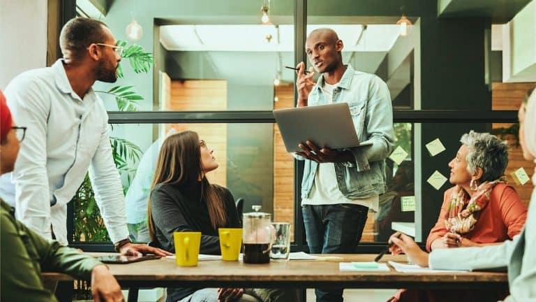 Group of work colleagues around a table looking at someone holding a laptop and talking